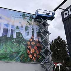 Fassadenwerbung Montage: Hubarbeitsbühne vor großer Werbefläche mit tropischem Motiv. Rundrohr-Konstruktion sichtbar.