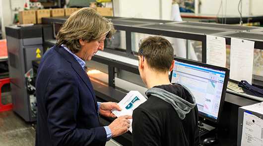 Downloads: Zwei Personen an einem Computer und einer Druckmaschine.