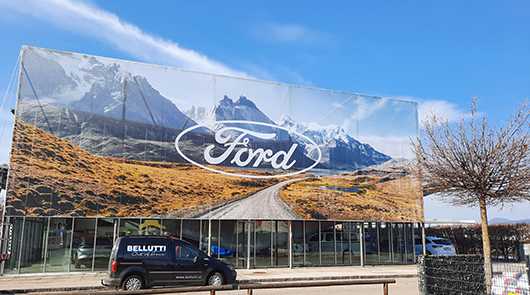 Außenwerbung: Ford-Banner mit Berglandschaft. Autohaus mit Fahrzeugen.